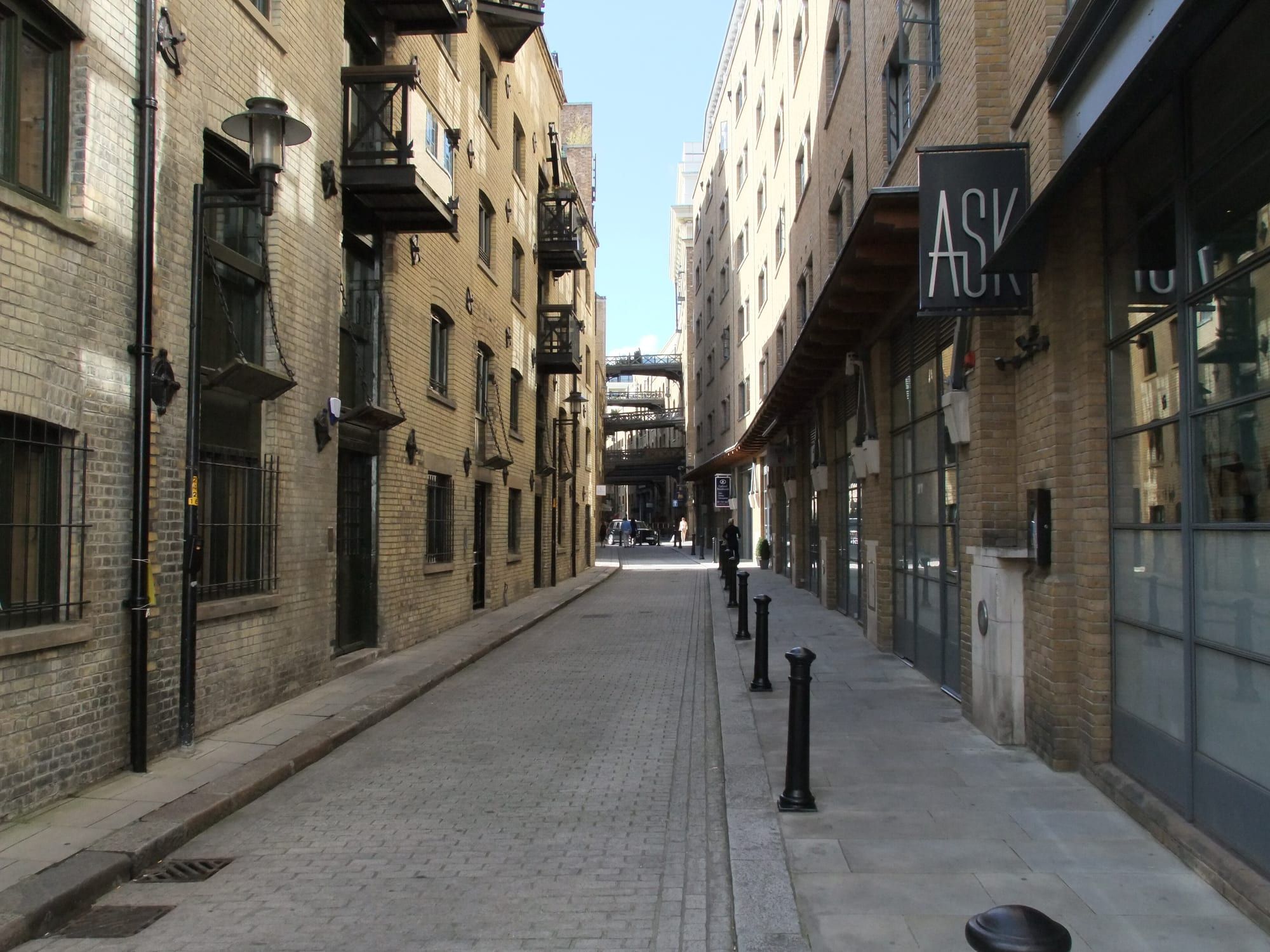 A narrow cobbled street between converted warehouses, with iron walkways in the distance